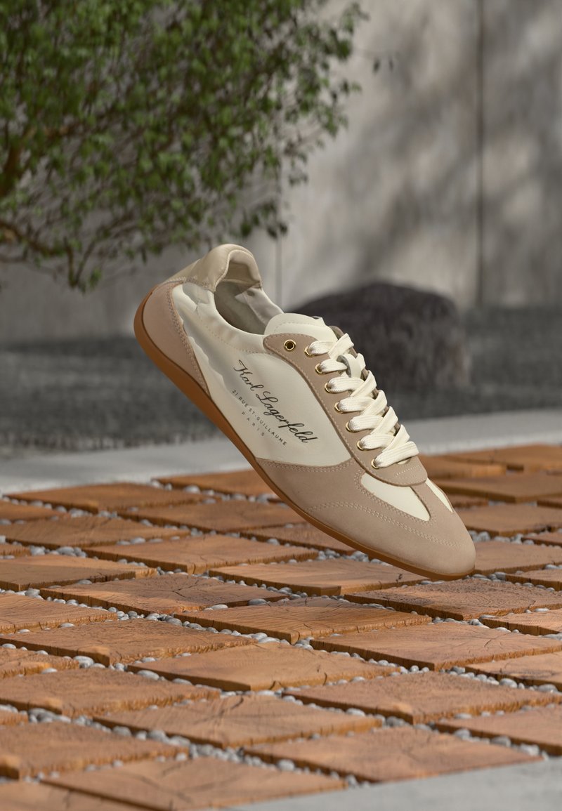 Μπέζ και λευκό αθλητικό παπούτσι Karl Lagerfeld με λευκά κορδόνια, αιωρούμενο πάνω από ξύλινη πλακόστρωτη επιφάνεια σε εξωτερικό χώρο και με θολό φόντο πράσινων φυτών.