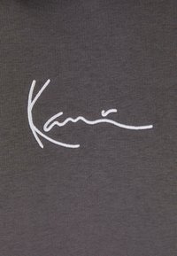 Grått stoff med hvit brodert signatur "Kami" i en flytende, kursiv design, som fremhever en jevn tekstur og minimalistisk stil.