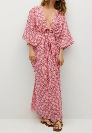 Kobieta ubrana w długą sukienkę w różowo-białe kwiaty z szerokimi rękawami, głębokim dekoltem w serek i wiązaniem w talii, w połączeniu z brązowymi sandałami.