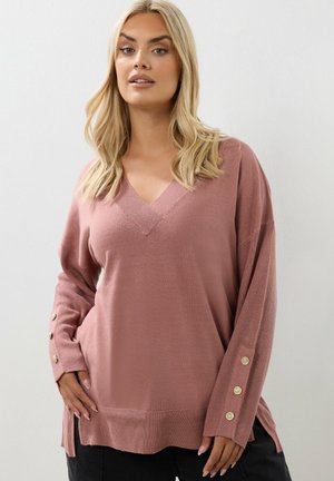 Luźny, różowy sweter dzianinowy z dekoltem w kształcie V, długimi rękawami i guzikami na mankietach. Posiada ribbowany dół oraz boczne rozcięcia.