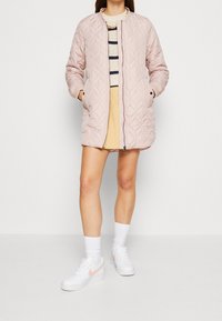 Lyserød, quilted lang jakke med rund halsudskæring, sidelommer og fuld lynlås, parret med en stribede sweater og en beige nederdel.