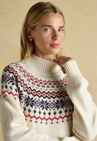 Pletený sveter má vysoký hrubý golier, krémovú farbu a viacfarebné geometrické vzory v červenej, modrej a hnedej farbe na hrudi.