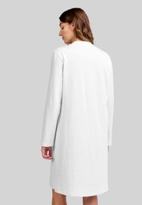 Mujer con cabello castaño hasta los hombros, vistiendo un vestido blanco sencillo de manga larga y hasta la rodilla, de pie contra un fondo gris claro.