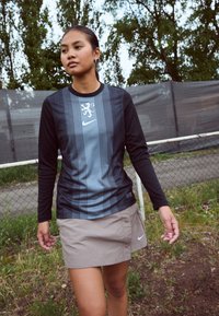 Nike long-sleeve shirt with black sleeves, striped front, and lion emblem; paired with a light brown skirt featuring buttons and logo.