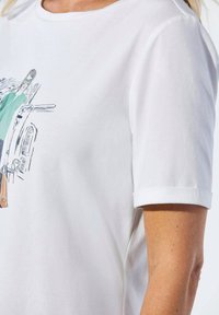 Weißes T-Shirt mit kurzen Ärmeln, das eine farbenfrohe Illustration einer Frau in einem grünen Kleid mit Sonnenbrille zeigt, die eine braune Tasche hält. Weicher Baumwollstoff.