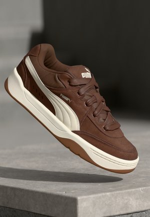 Brązowy sneaker Puma z białym paskiem i podeszwą unoszący się nad szarą betonową powierzchnią na ciemnym tle.