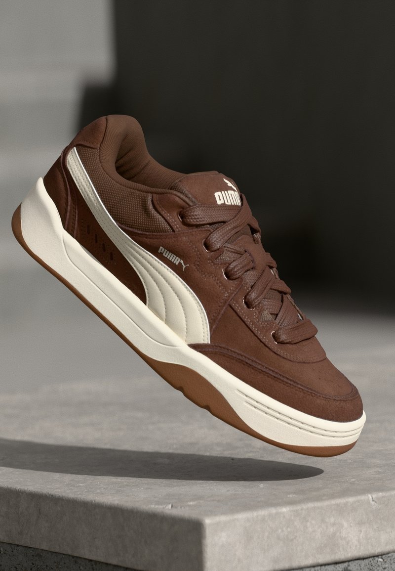 Baskets Puma marron avec bande blanche et semelle flottante au-dessus d'une surface en béton grise sur fond sombre.