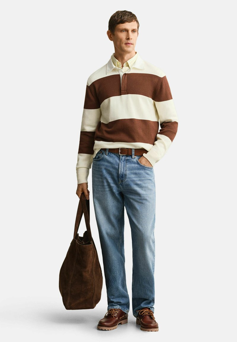 Mann trägt braunen und weißen gestreiften Pullover, hellblaue Jeans, braune Schuhe, hält eine große braune Wildleder-Tragetasche.