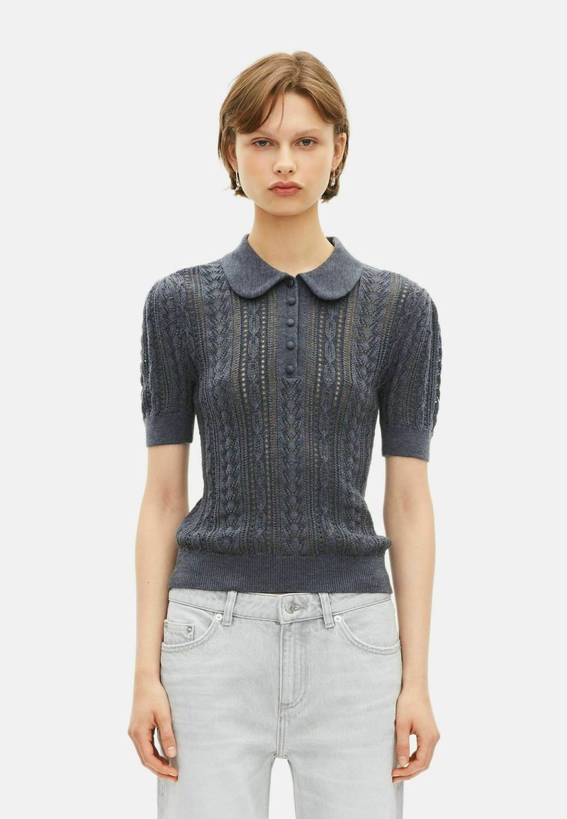 Młoda osoba w ciemnoniebieskim, krótkim swetrze z dzianiny z kołnierzykiem typu Peter Pan i jasnoszarych dżinsach, stojąca na jednolitym tle.