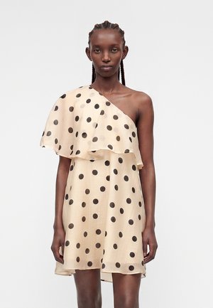 Femme avec des tresses portant une robe beige à une épaule avec des pois noirs, debout sur un fond blanc uni.