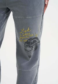 Grå bomullsshorts med en broderad tigergrafik och frasen "Tur är där förberedelse möter möjlighet" i gult.