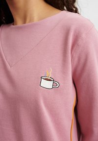 Rosa sweatshirt med en V-ringning. Har en broderad kaffekopp med ånga. Inkluderar detaljer med orange sidostråk. Mjuk, texturerad tyg.