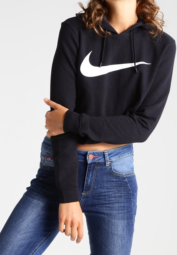 Černá zkrácená mikina s velkým bílým logem Nike swoosh, v kombinaci s modrými džíny. Tkanina vypadá měkká s hladkým povrchem.