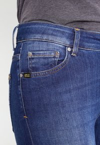 Jeanbyxor i mörkblått, med subtila blekningar, gul-orange sömnad och en liten märkesetikett på framfickan.