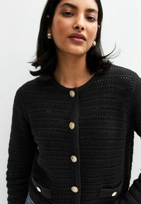 Cardigan en tricot noir avec un motif texturé, doté de boutons dorés et de poches. Le design a un col rond et des manches longues.