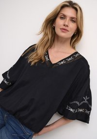 Top nero con scollatura a V, dettagli in pizzo trasparente e modelli floreali ricamati sulle maniche. Vestibilità ampia e texture liscia. Indossato con jeans blu.