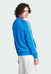 Casaco de pista azul com riscas brancas. Tecido suave, fecho de correr completo, punhos canelados, colarinho alto. Usado sobre calças brancas, visto de costas.