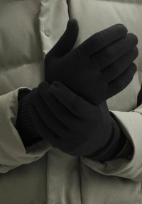 Czarne, dziergane rękawiczki o prążkowanej strukturze. Ręce złożone razem na tle szarej, pikowanej kurtki. Tkanina wydaje się miękka i elastyczna.