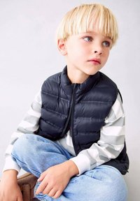 Čierna vypchávkovaná vesta cez šedé a biele pruhované tričko s dlhým rukávom. Svetlomodré džínsové nohavice. Dieťa sedí s neutrálnym výrazom.
