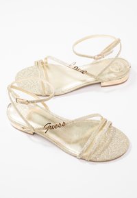 Paire de sandales plates dorées pailletées avec des brides à la cheville, des fines brides avant et des détails métalliques "Guess" et "Love" sur les semelles.