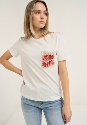 Mujer con camiseta blanca de manga corta con un bolsillo texturizado adornado con decoraciones florales rojas y rosas, combinada con vaqueros azul claro.