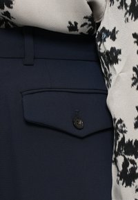 Granatowe spodnie z boczną kieszenią z czarnym guzikiem, w połączeniu z wzorzystą bluzką z lekkiego materiału w odcieniach beżu i czerni.