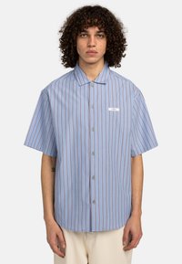 Camicia a maniche corte con bottoni in tessuto azzurro chiaro, con righe verticali rosse e bianche, dotata di colletto a punta e di una piccola etichetta con logo.