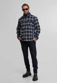 Giacca di flanella a scacchi in blu navy e bianco, con colletto, due tasche sul petto, chiusura a bottoni e vestibilità comoda, indossata con pantaloni scuri e stivali.