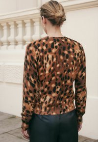 Une femme aux cheveux blonds en chignon porte un pull en imprimé léopard marron et noir ainsi que des pantalons en cuir noir, se tenant à l'extérieur près d'une balustrade en pierre.