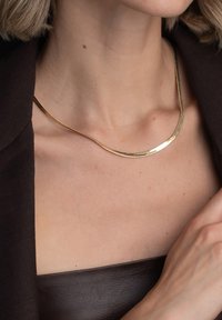 Guldtonad halsband med en böjd, polerad design. Slät textur, minimalistisk form, sitter runt halsen. Ingen synlig spänne eller prydnader.