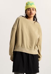 Felpa beige con vestibilità oversize, colletto e orlo a coste, indossata sopra una tunica nera. Il modello indossa un berretto giallo brillante con un design verde.