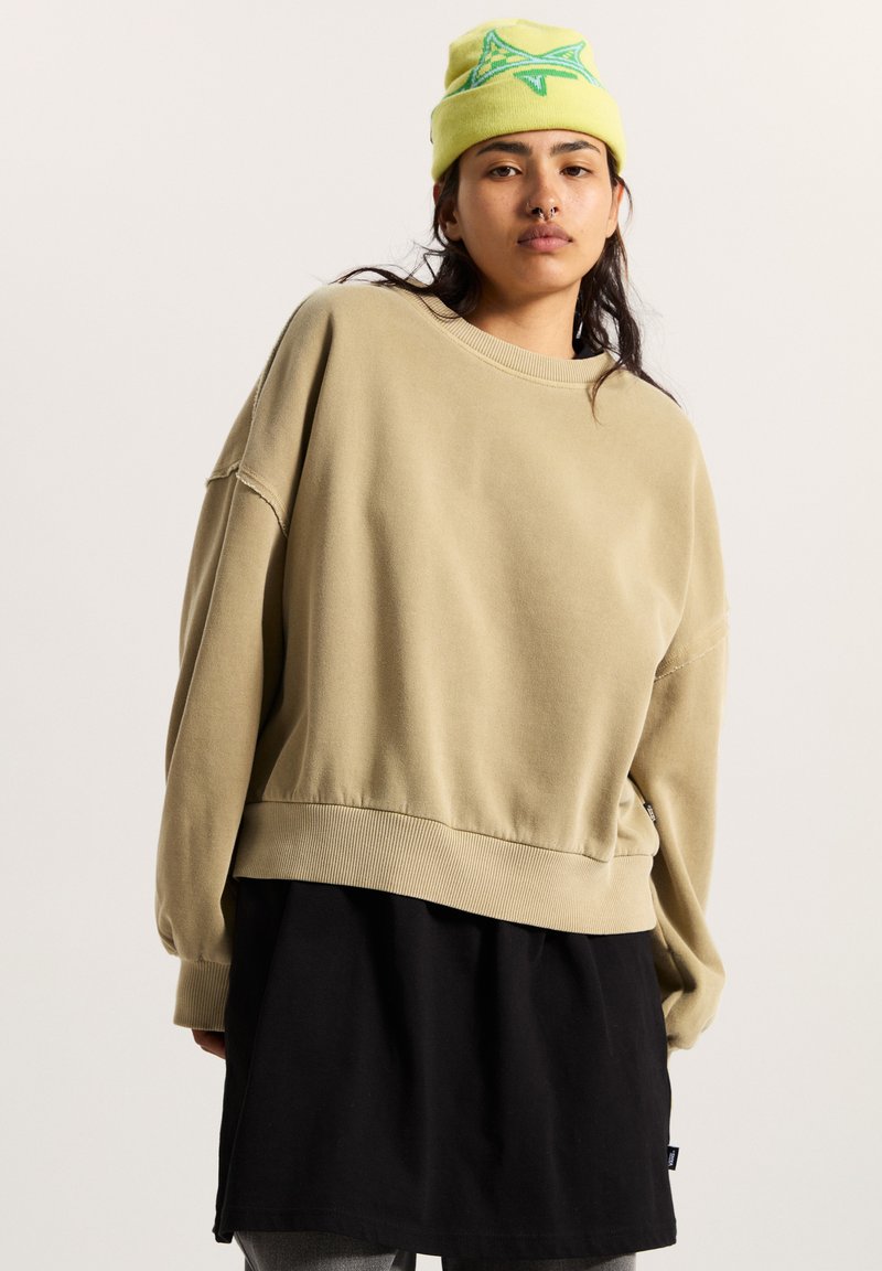 Felpa beige con vestibilità oversize, colletto e orlo a coste, indossata sopra una tunica nera. Il modello indossa un berretto giallo brillante con un design verde.