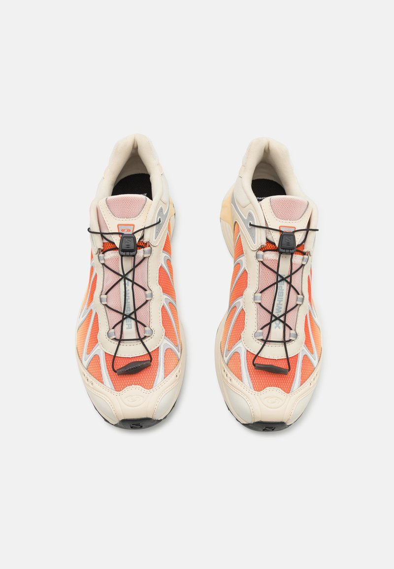 Zapatillas con una parte superior de malla en beige y naranja, cordones negros y una suela de goma con un acabado texturizado. Diseño moderno con detalles en contraste.