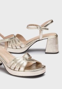 Sandales métalliques dorées à bout ouvert, avec un design noué sur le devant, une bride de cheville et un talon moyen.