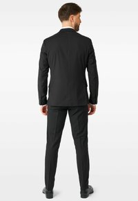 Traje negro con corte ajustado, chaqueta de un solo botón y pantalones de frente plano. Presenta detalles sutiles en los bolsillos y una tela texturizada.