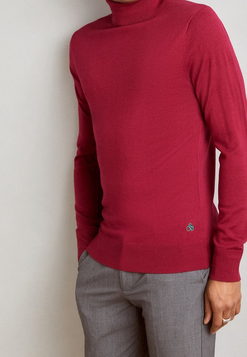 Homme portant un pull à col roulé rouge ajusté associé à un pantalon gris, la main posée près de la poche sur un fond uni.