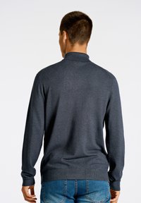 Mörkgrå pullovertröja med hög krage, lång ärm och ribbade cuffs. Bärs tillsammans med blå denimjeans som visar en slät tygtextur.