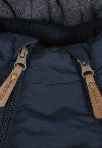 Jachetă izolată de culoare bleumarin, cu închidere prin fermoar dotat cu mânere din piele maro, manșete ribbate și o căptușeală interioară moale de culoare gri pentru un plus de căldură.