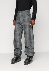 Moški v sivih karo snežnih hlačah z zadrgo na stranski žep in črnimi smučarskimi čevlji, ki stoji pred enostavno svetlo ozadjem.