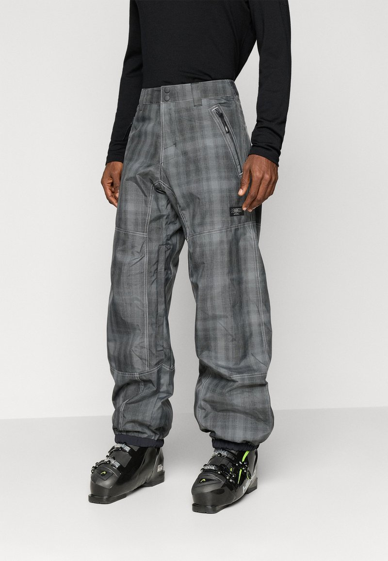 Moški v sivih karo snežnih hlačah z zadrgo na stranski žep in črnimi smučarskimi čevlji, ki stoji pred enostavno svetlo ozadjem.