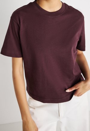 Osoba nosząca gładki ciemnofioletowy krótki rękaw T-shirt i białe spodnie, z jedną ręką w kieszeni.