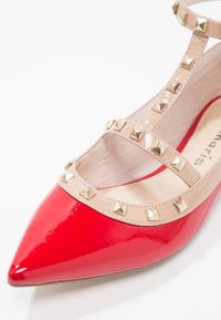 Chaussure en cuir verni rouge avec un bout pointu et des brides de cheville couleur nude ornées de clous pyramides dorés. Texture lisse et design minimaliste.