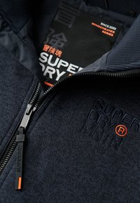 Veste en polaire marine à finitions texturées, fermeture éclair et logo brodé. Comprend une étiquette indiquant "Superdry Windcheater Series."
