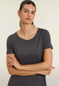 Camiseta gris con mangas cortas y cuello redondo. En la parte frontal presenta un diseño con tachuelas que dice "FREDDY". Textura de tela suave.