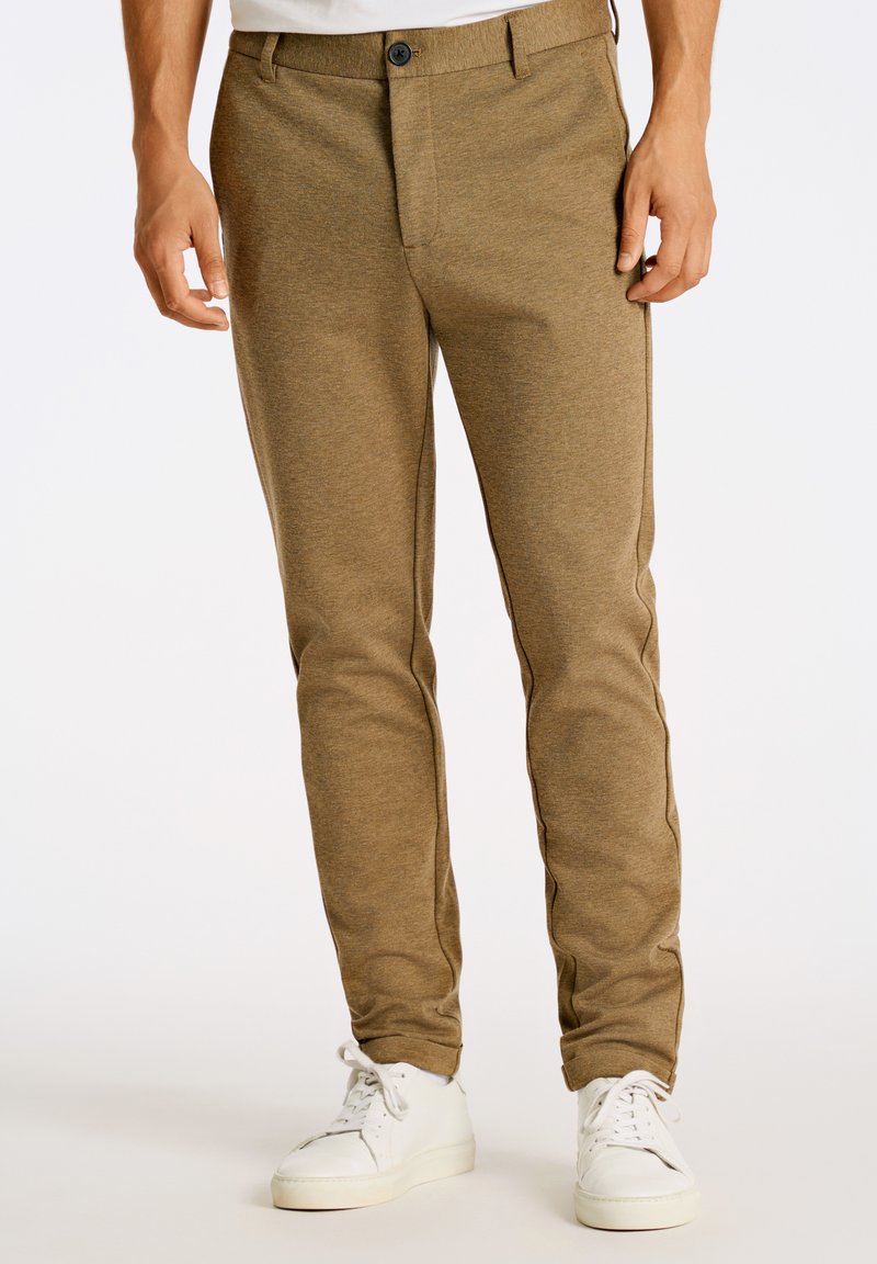Pantaloni marroni con texture e vestibilità slim, chiusura con bottone e tasche; abbinati a scarpe da ginnastica bianche per un look casual.