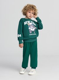 Felpa in cotone verde e pantaloni abbinati, con una grafica di un gorilla dei cartoni animati, con dettagli in bianco tra testo e numeri. Scarpe da ginnastica bianche.