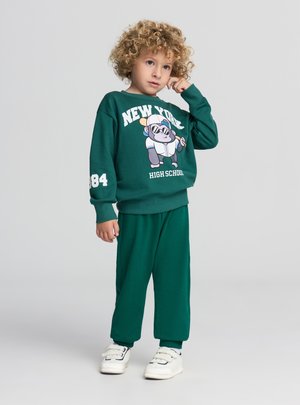 Felpa in cotone verde e pantaloni abbinati, con una grafica di un gorilla dei cartoni animati, con dettagli in bianco tra testo e numeri. Scarpe da ginnastica bianche.