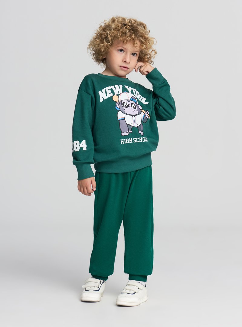 Felpa in cotone verde e pantaloni abbinati, con una grafica di un gorilla dei cartoni animati, con dettagli in bianco tra testo e numeri. Scarpe da ginnastica bianche.