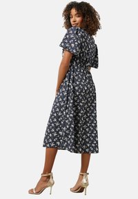 Vestido floral azul marino con mangas cortas abullonadas, cintura ajustada y falda fluida. Combinado con sandalias de tacón alto metálicas doradas.