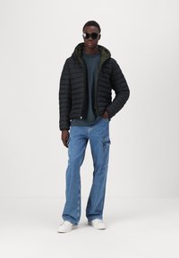Doudoune noire avec un intérieur vert, pantalon cargo en denim bleu et baskets blanches. Le modèle porte des lunettes de soleil et se tient devant un fond uni.